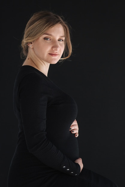 Feche o retrato de uma linda mulher grávida em um vestido preto, segurando a mão dela na barriga e olhando para baixo