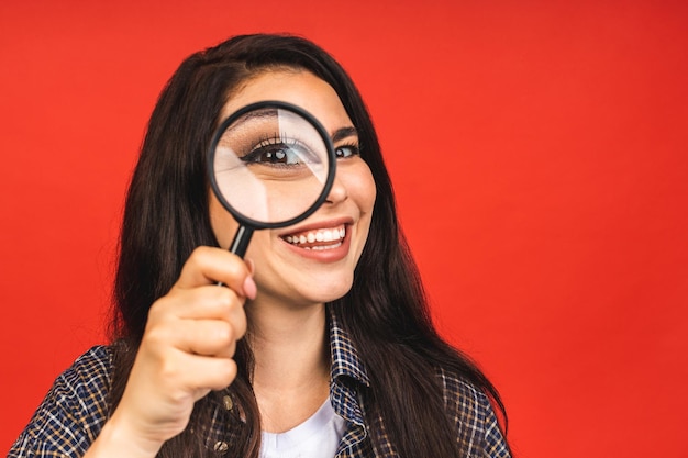 Feche o retrato de uma linda mulher incrível olhando para a câmera através de uma lupa isolada em fundo vermelho se divertindo