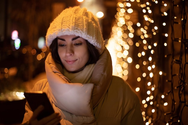 Feche o retrato de uma linda mulher sorridente com chapéu de inverno branco e jaqueta enquanto ela segura e usa seu telefone inteligente durante a caminhada ao ar livre com iluminação de natal da cidade no fundo