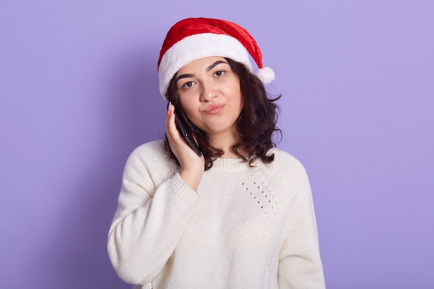 Feche o retrato de uma menina linda morena com chapéu de papai noel falando no celular isolado sobre uma parede lilás