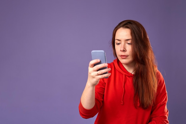 Feche o retrato de uma mulher caucasiana em roupas vermelhas usando seu celular em fundo roxo