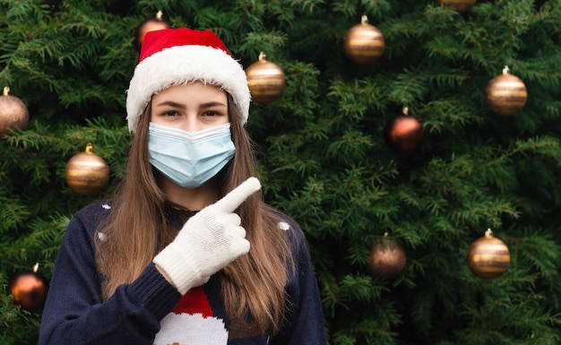 Feche o retrato de uma mulher com um chapéu de Papai Noel e uma máscara médica com emoção e mostre o dedo. No contexto de uma árvore de Natal. Pandemia do coronavírus