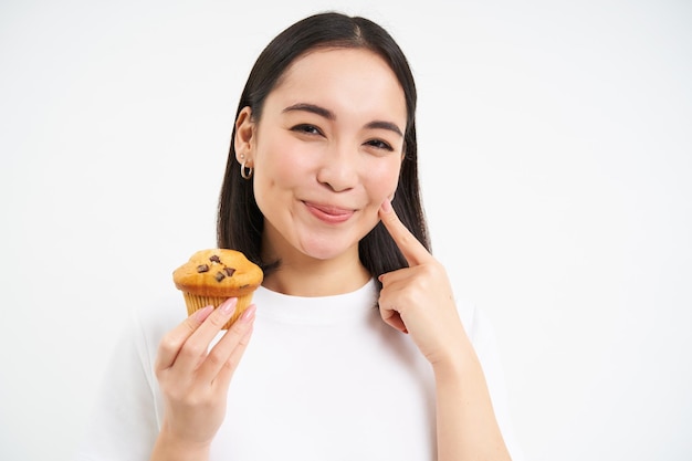 Feche o retrato de uma mulher coreana sorridente comendo cupcake com fundo branco de rosto satisfeito