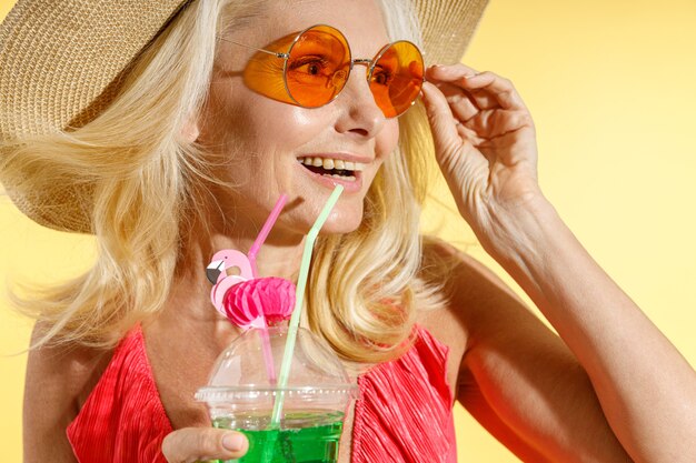 Feche o retrato de uma mulher loira alegre em óculos de sol, maiô vermelho e chapéu de palha sorrindo à parte