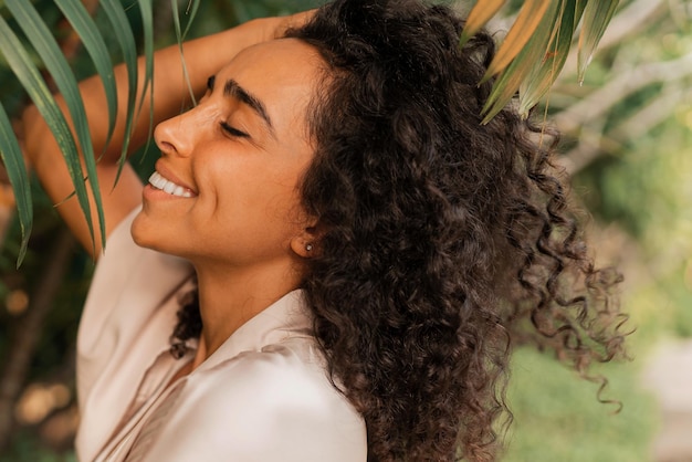 Feche o retrato de uma mulher rindo com cabelos ondulados posando no jardim tropical. Vestindo roupão de seda elegante. Conceito de spa e bem-estar.