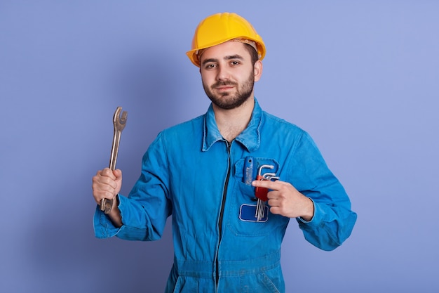 Feche o retrato do engenheiro barbudo com chaves, olhando para a câmera e apontando para sua ferramenta com o dedo indicador, reparador posando contra azul, mecânico veste uniforme e capacete amarelo.