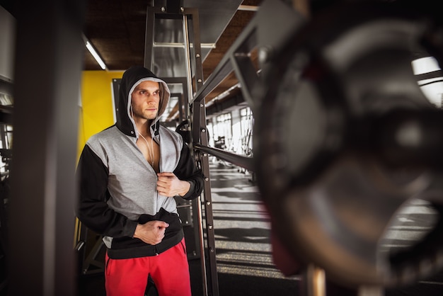 Feche o retrato do homem encapuzado muscular focado e motivado em pé com fones de ouvido no ginásio moderno.