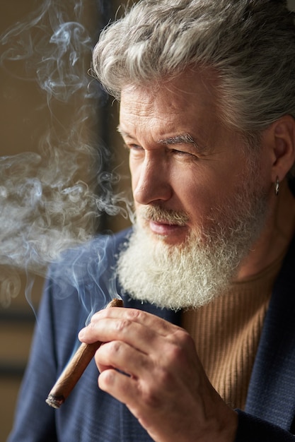 Feche o retrato do homem maduro de cabelos grisalhos sério, olhando para longe enquanto fumava charuto. Estilo de vida, sucesso, conceito de pessoas
