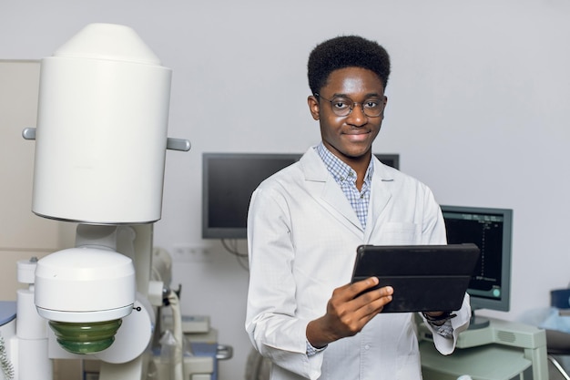 Feche o retrato do jovem médico afro-americano em uniforme branco e óculos segurando o tablet digital digital posando perto da moderna máquina de litotriptor ultrassônico no centro médico de urologia