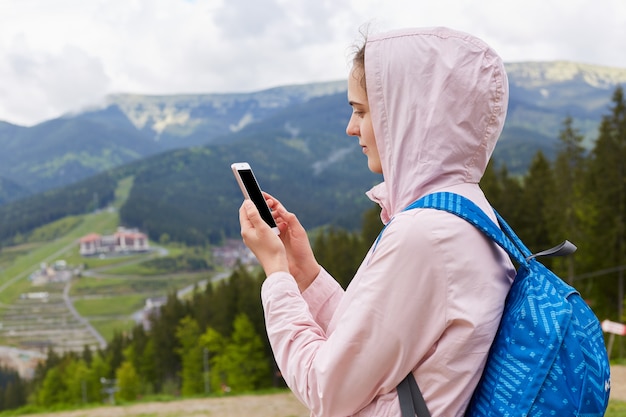 Feche o retrato do perfil de mulher jovem alpinista com mochila azul usando smartphone na bela paisagem da montanha na primavera