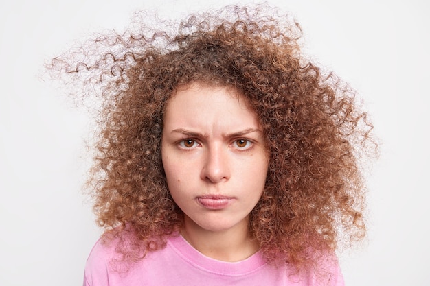 Feche o retrato do rosto carrancudo de sorrisos de mulher de cabelo encaracolado mal-humorado ofendeu satres de expressão suspeita conturbada com insatisfação isolada sobre uma parede branca. Conceito de emoções.