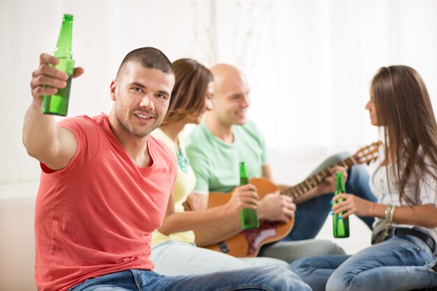 Feche-se de um jovem sorrindo em casa festa com cerveja e aplausos. seus amigos ao fundo.