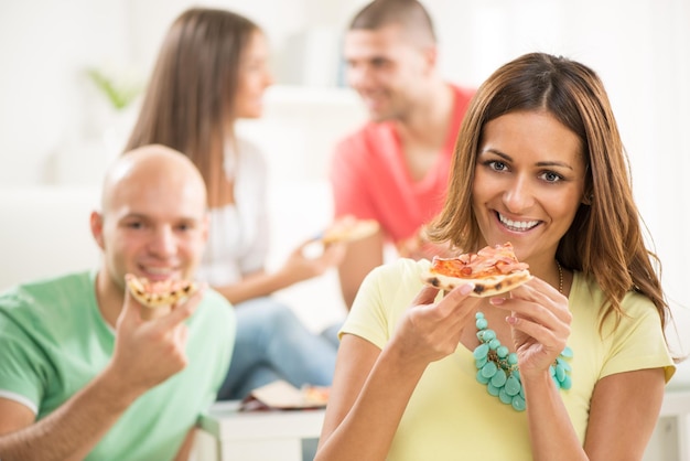 Feche-se de uma jovem sorrindo e comendo pizza com seus amigos ao fundo.