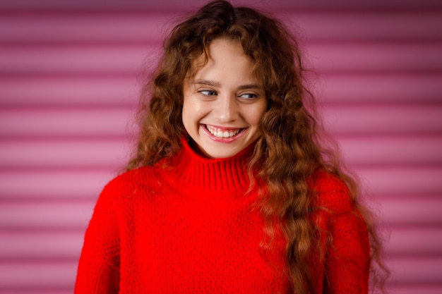 Foto feche uma jovem adulta sorridente usando um suéter vermelho de inverno dentro de casa, olhando para a câmera com um sorriso alegre