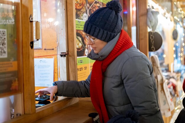 Feenmarkt in Nokolskie ryady. Kaukasische Frau mit Hut und rotem Schal, die Leckereien im Kiosk kauft. Kontaktloses Bezahlen auf dem Weihnachtsmarkt. Bild mit selektivem Fokus und Rauscheffekt