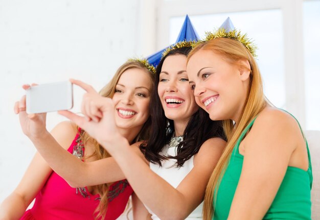 Feier, Freunde, Junggesellenabschied, Geburtstagskonzept - drei lächelnde Frauen in blauen Hüten, die Spaß mit der Smartphone-Fotokamera haben
