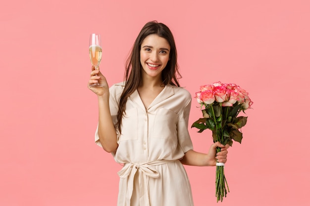 Feier-, Jubel- und Schönheitskonzept. Schöne junge zarte Frau im Kleid, die Blumenstraußblumen hält, Glas Champagner erhebt, Toast gibt, lächelt und Party, rosa Wand genießt