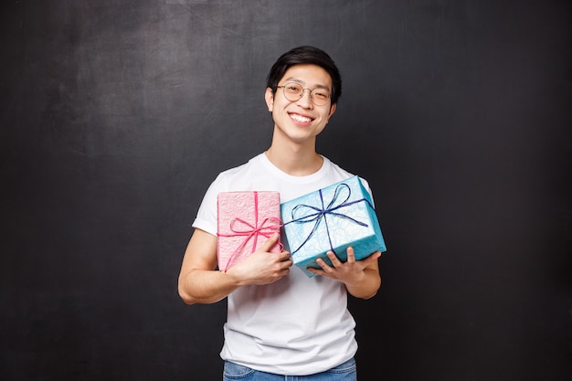 Feier, Urlaub und Lifestyle-Konzept. Porträt des niedlichen und gutaussehenden Freundes bereitete Geschenke für seinen Partner vor, der zwei Geschenkboxen rosa und blau hält und stolz, schwarze Wand lächelt