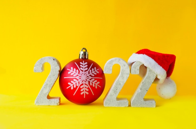 Feiern des neuen Jahres 2022. Nummer 2022 mit konkreten Ziffern in Weihnachtsmütze und Weihnachtskugel auf gelbem Hintergrund.