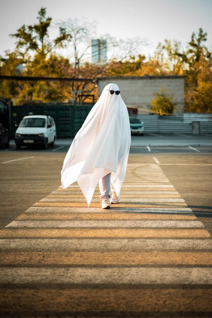 Feiern Sie Halloween Ghost Challenge 2021 Ein Mann, der als Geist aus einem Laken und Sonnenbrille gekleidet ist, überquert die Straße entlang der Fußgängerüberfahrt.