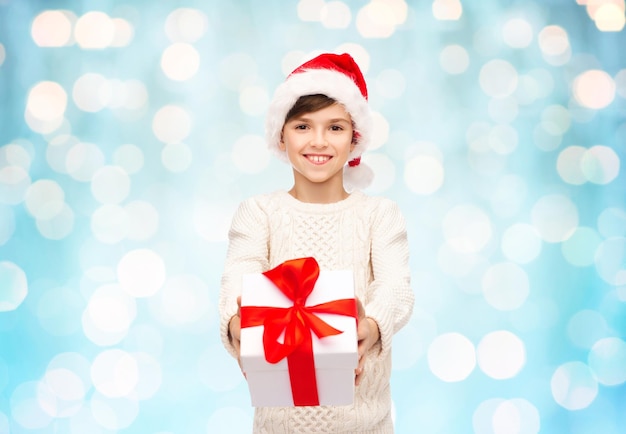 feiertage, geschenke, weihnachten, kindheit und menschenkonzept - lächelnder glücklicher junge in weihnachtsmütze mit geschenkbox über blaulichthintergrund