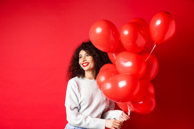 Feiertage und Feier. Glückliche Frau, die mit Partyballons auf roter Wand aufwirft, den leeren Raum beiseite schauend.