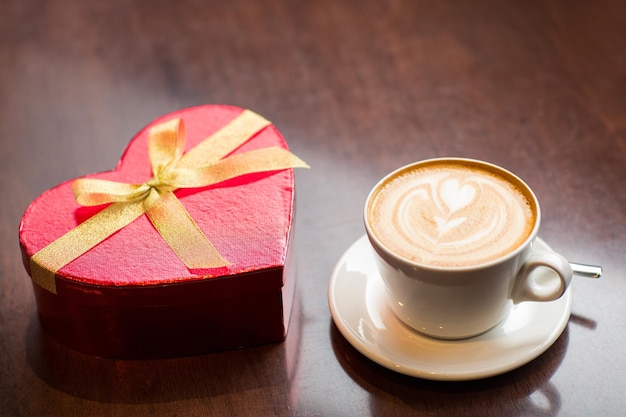 Feiertage, Valentinstag, Liebe und Getränkekonzept - Nahaufnahme von Geschenkbox und Kaffeetasse auf dem Tisch