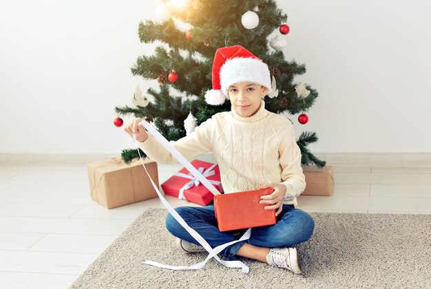 Feiertage, Weihnachten, Kindheit und Leutekonzept - lächelnder glücklicher jugendlich Junge in Sankt-Hut öffnet Geschenkbox über Weihnachtsbaumhintergrund.