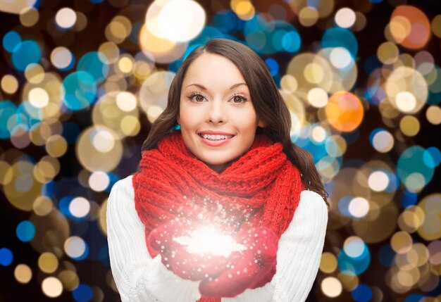 Foto feiertage, weihnachten und menschen konzept - glückliche frau in schal und handschuhe mit magischer schneeflocke über lichtern hintergrund