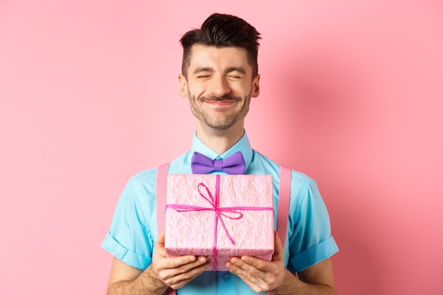 Feiertags- und Feierkonzept. Glücklicher Kerl mit französischem Schnurrbart, der vor Freude grinst, niedliche Geschenkbox zeigt, Geschenk empfangend, über rosa Hintergrund stehend.
