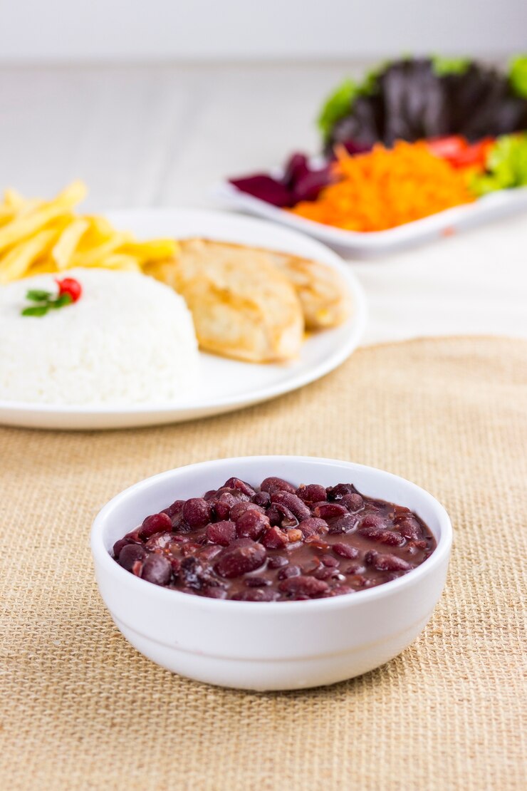 Foto feijão preto em prato branco em pano de prato de algodão cru com um prato cheio com arroz, frango e batatas fritas atrás