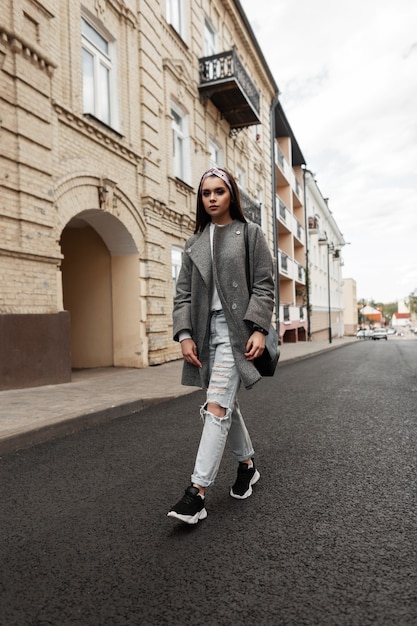 Feine hübsche junge frau im glamour-bandana mit schwarzer tasche im grauen mantel mit handtasche in schuhen geht in der nähe des vintage-gebäudes auf der straße. nettes mädchenmodell reisen auf der straße in der stadt. stylisches frühlings-outfit für damen.