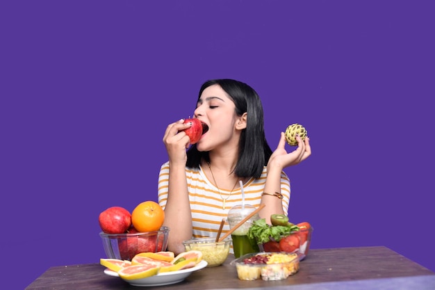 Feinschmeckermädchen, das am Obsttisch sitzt und Apfel über lila Hintergrund isst, indisches pakistanisches Modell?