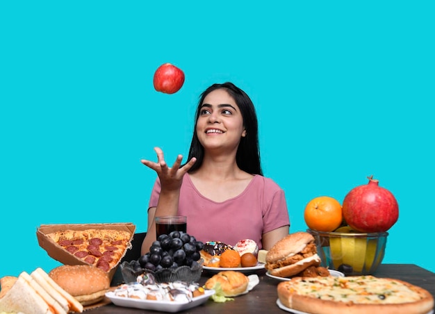 Feinschmeckermädchen, das am Obsttisch sitzt und lächelt und rote Äpfel fängt, indisches pakistanisches Modell?
