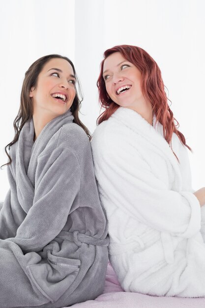 Felices amigas en batas de baño sentado espalda con espalda en la cama