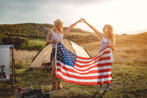 Felices amigas jóvenes disfrutan de un día soleado en la naturaleza. Sostienen una bandera estadounidense frente a una tienda de campaña.