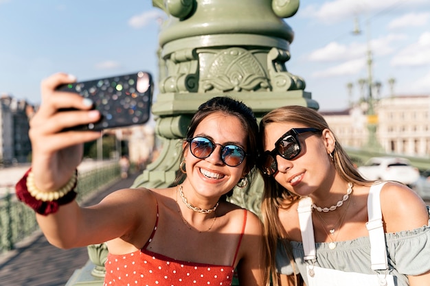 Foto felices amigas jóvenes en gafas de sol sentado cerca de la columna y navegar por internet en el teléfono móvil de la calle de la ciudad