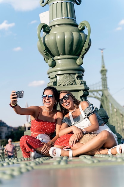 Felices amigas jóvenes en gafas de sol sentado cerca de la columna y navegar por internet en el teléfono móvil de la calle de la ciudad