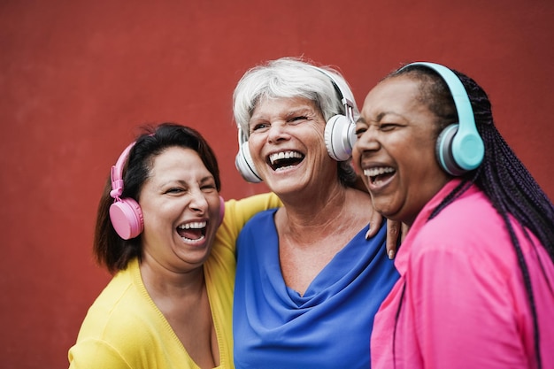 Felices amigas maduras divirtiéndose juntas mientras escuchan música con auriculares inalámbricos Las mujeres multiétnicas disfrutan del día al aire libre