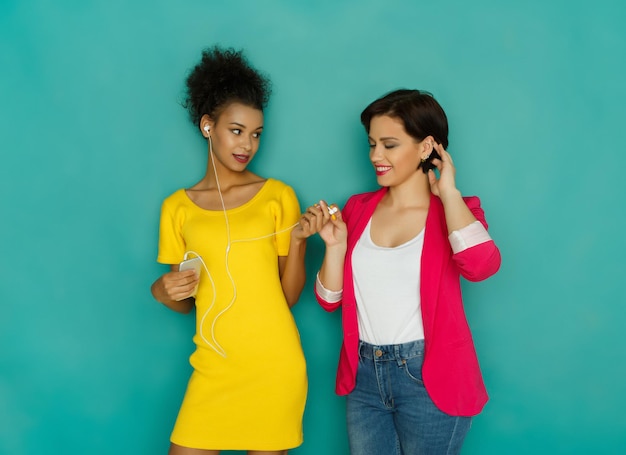 Felices amigas multiétnicas. Chica negra compartiendo audífonos con su amiga escuchando música en el teléfono inteligente en el fondo turquesa con espacio de copia