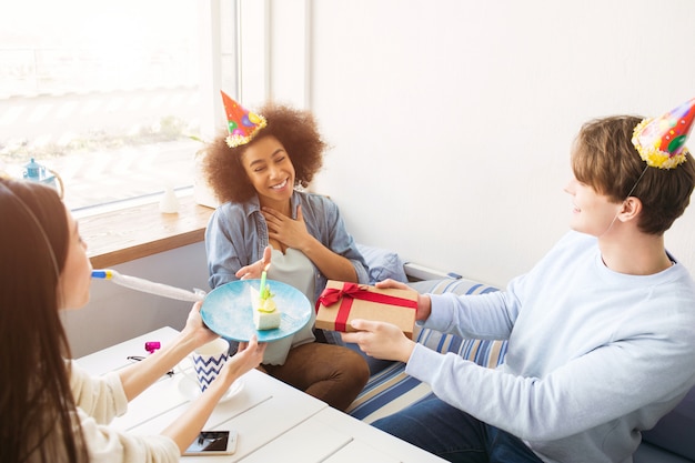Felices amigos celebran cumpleaños. Tienen sombreros divertidos en la cabeza. Guy sostiene un regalo mientras la chica del suéter blanco sostiene un plato con un trozo de pastel. Chica afroamericana es feliz.
