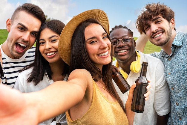 Felices amigos multirraciales se toman selfies mientras disfrutan de una cerveza al aire libre en la naturaleza mirando a la cámara