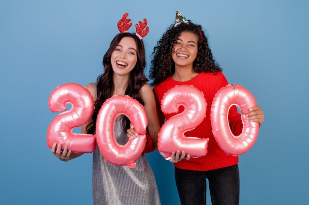 Felices chicas multirraciales con globos de año nuevo 2020 y divertidos aros festivos aislados en azul