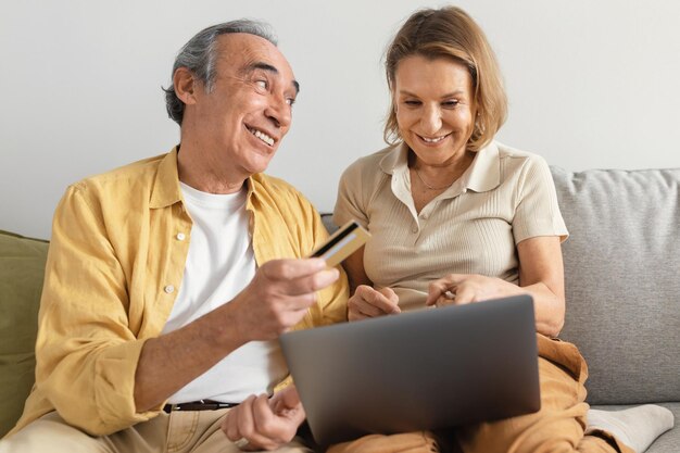 Felices cónyuges mayores comprando en línea en un portátil que le da tarjeta de crédito a su esposa sentada en un sofá en casa
