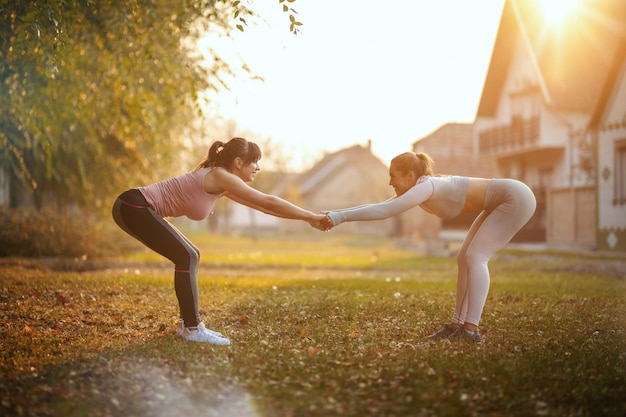 Felices dos amigas están haciendo ejercicios de estiramiento en la naturaleza en un día soleado.