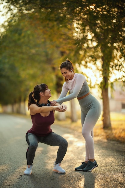 Foto felices dos mujeres jóvenes están haciendo ejercicios de calentamiento antes de correr por el soleado sendero de la avenida.