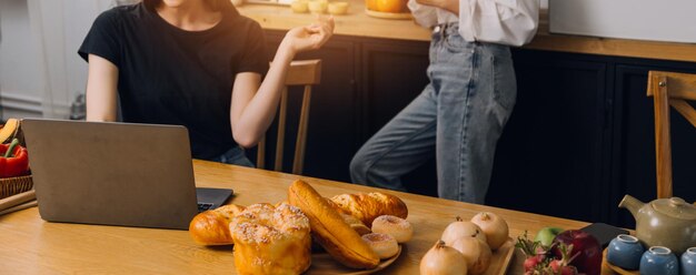 Felices dos mujeres jóvenes que buscan una computadora portátil mientras cocinan juntas en la cocina de su casa Dos jóvenes lesbianas diversas que pasan tiempo juntas LGBT y el concepto de identidad de género