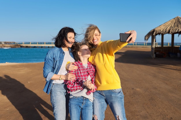 Felices dos mujeres de mediana edad y una niña tomando selfie en el fondo de la playa del mar del teléfono inteligente