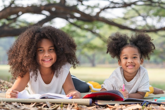 Felices dos niñas afroamericanas mintiendo dibujando en el libro de colorear para niños en el parque