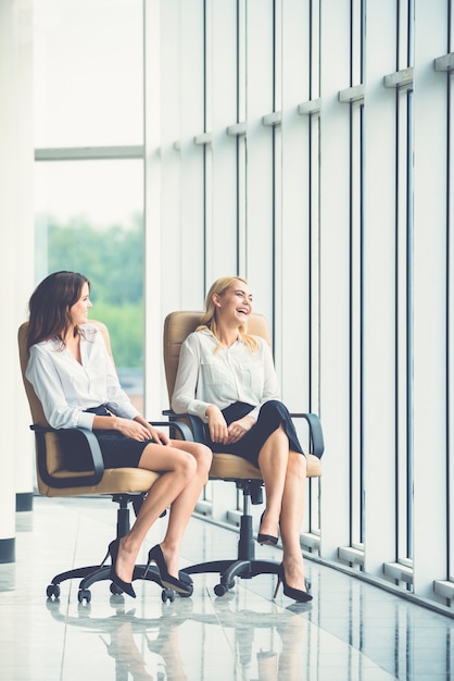 Las felices empresarias en sillas hablan cerca de la ventana de la oficina.